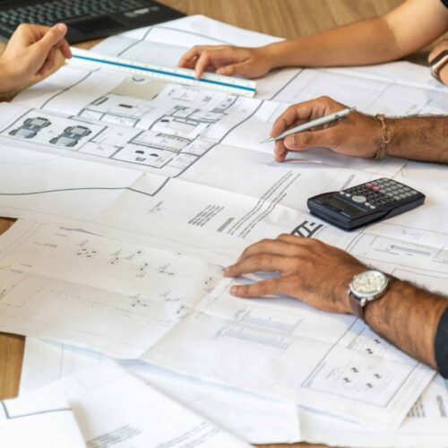pedro-miranda-3QzMBrvCeyQ-unsplash (1) Twee personen bekijken architecturale bouwplannen op tafel met rekenmachine en laptop tijdens een renovatieadviesgesprek